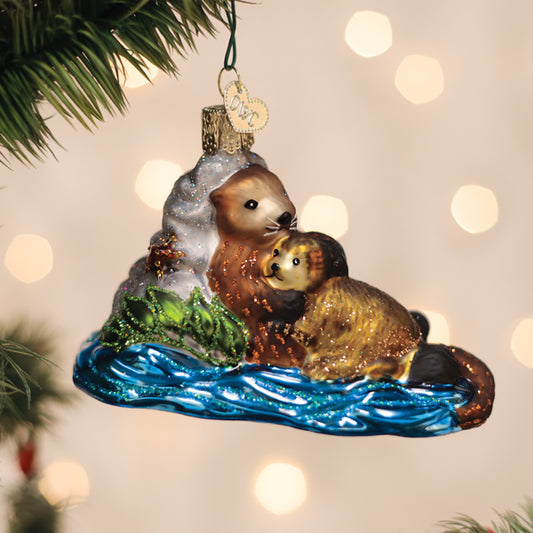 A glass Christmas tree ornament showing two otters cuddling on blue water, with one otter resting its head on the other. The ornament hangs from a green branch, with soft lights blurred in the background.