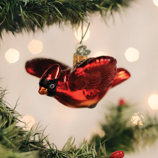 A shiny red cardinal bird ornament hangs on a green Christmas tree branch, with soft, out-of-focus lights glowing in the background.
