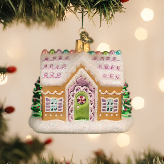 A gingerbread house ornament with snowy details and pastel colors hangs on a Christmas tree, surrounded by blurred festive lights in the background.
