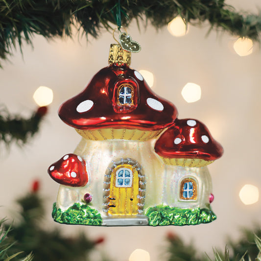 A glass Christmas tree ornament shaped like a whimsical mushroom house, with red caps and white spots, hangs from a tree branch with blurred lights in the background.