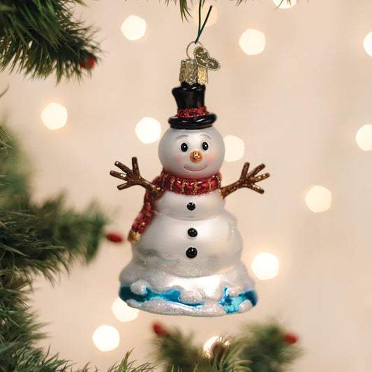 A glass snowman ornament with a red scarf, black buttons, and a top hat hangs on a Christmas tree branch. Warm white lights softly glow in the blurred background.