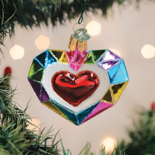 A colorful, heart-shaped Christmas ornament with a shiny red heart in the center hangs on a green tree branch, with blurred festive lights in the background.