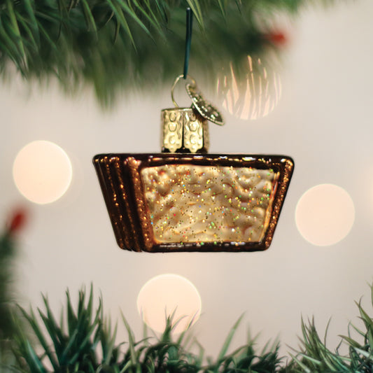 A glittery ornament shaped like a golden panettone cake hangs from a Christmas tree branch, with blurred lights glowing softly in the background.