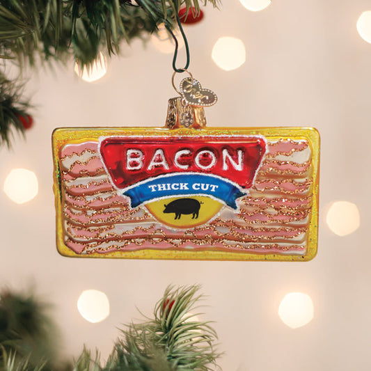 A Christmas tree ornament shaped like a package of thick cut bacon hangs on a tree branch, with festive lights softly glowing in the background.