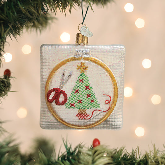 A square Christmas tree ornament with an embroidered design of a decorated tree, red scissors, and thread, hanging on a green branch with sparkling lights in the background.