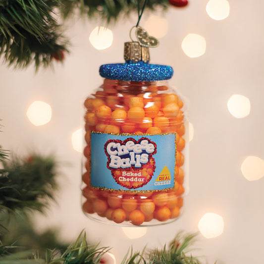 A Christmas tree ornament shaped like a plastic jar filled with orange cheese balls hangs from a branch. The jar has a blue glittery lid and a label reading Cheese Balls, Baked Cheddar. Blurred holiday lights are in the background.