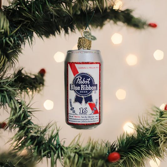 A Christmas tree branch decorated with a Pabst Blue Ribbon beer can ornament, surrounded by green needles and small red berries, with blurred warm lights in the background.