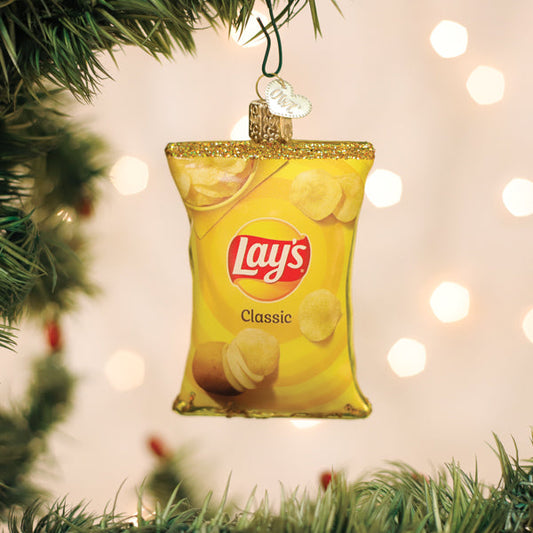 A Christmas tree ornament shaped like a yellow Lays Classic potato chips bag hangs from a branch, surrounded by green pine needles and blurred festive lights in the background.