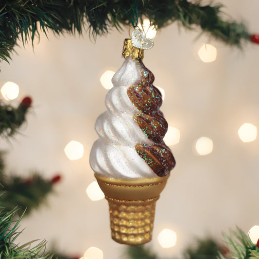 A Christmas tree ornament shaped like a glittery chocolate and vanilla swirl ice cream cone hangs from a green branch, with blurred lights in the background.