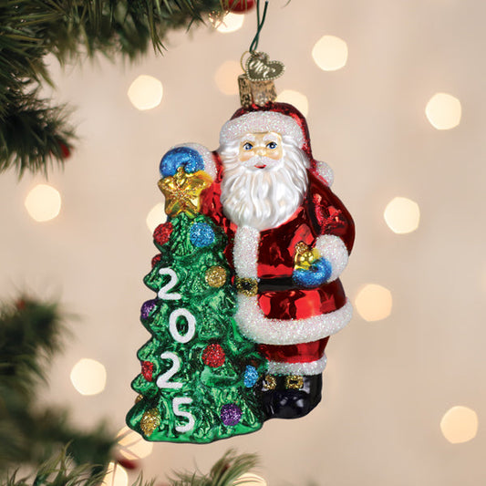 A festive glass ornament of Santa Claus holding a decorated Christmas tree with colorful ornaments and the year 2025 on it, hanging on a Christmas tree with blurred lights in the background.