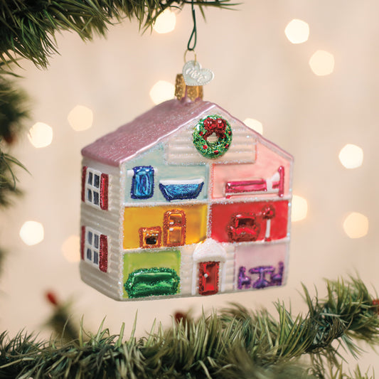 A colorful glass Christmas ornament shaped like a dollhouse hangs on a Christmas tree. The ornament shows rooms with furniture and a green wreath above the door, with blurred festive lights in the background.