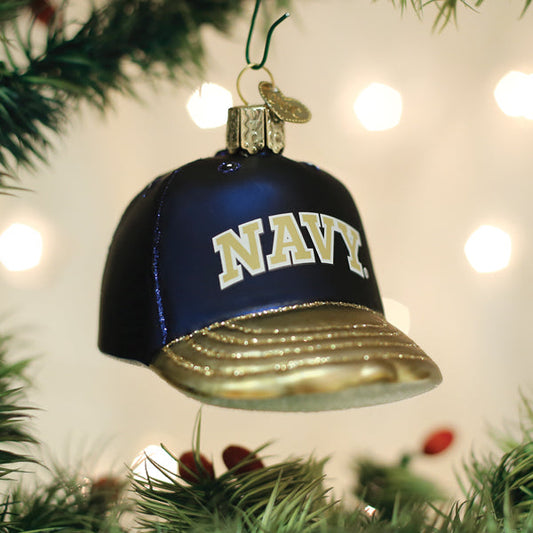 A Christmas tree ornament shaped like a navy blue and gold baseball cap with â€œNAVYâ€_x009d_ written on the front, hanging among green tree branches with blurred lights in the background.