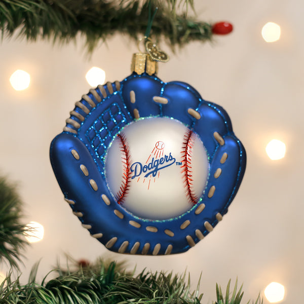 A Christmas tree ornament shaped like a blue baseball glove holding a baseball with the Dodgers logo, hanging on a tree with blurred white lights in the background.