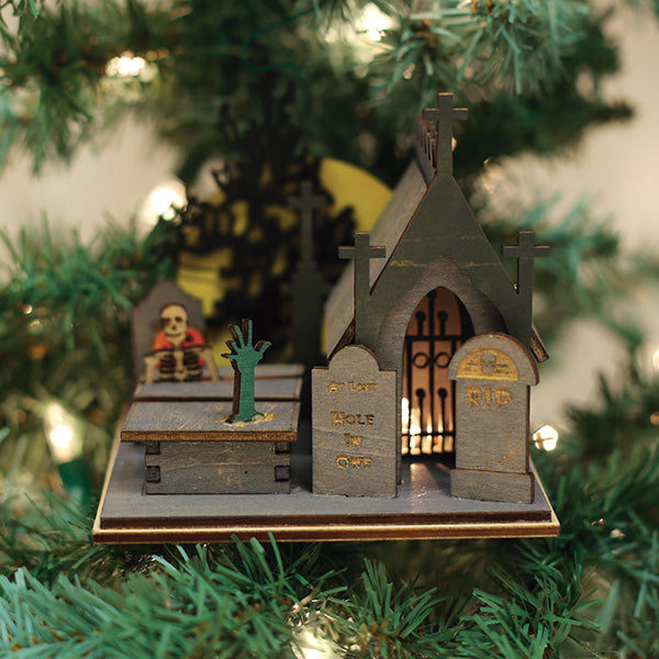 A spooky Halloween-themed ornament on a Christmas tree features a haunted church, tombstones, a skeleton, and a green hand rising from a grave, all surrounded by green pine needles and warm lights.