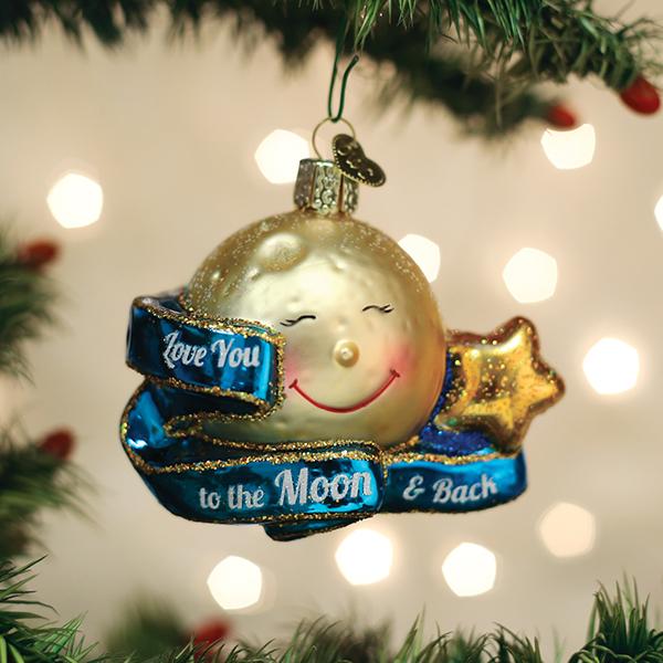 A gold, smiling moon ornament with a star and a blue ribbon that says Love you to the Moon & Back hangs from a Christmas tree branch, with twinkling lights in the background.