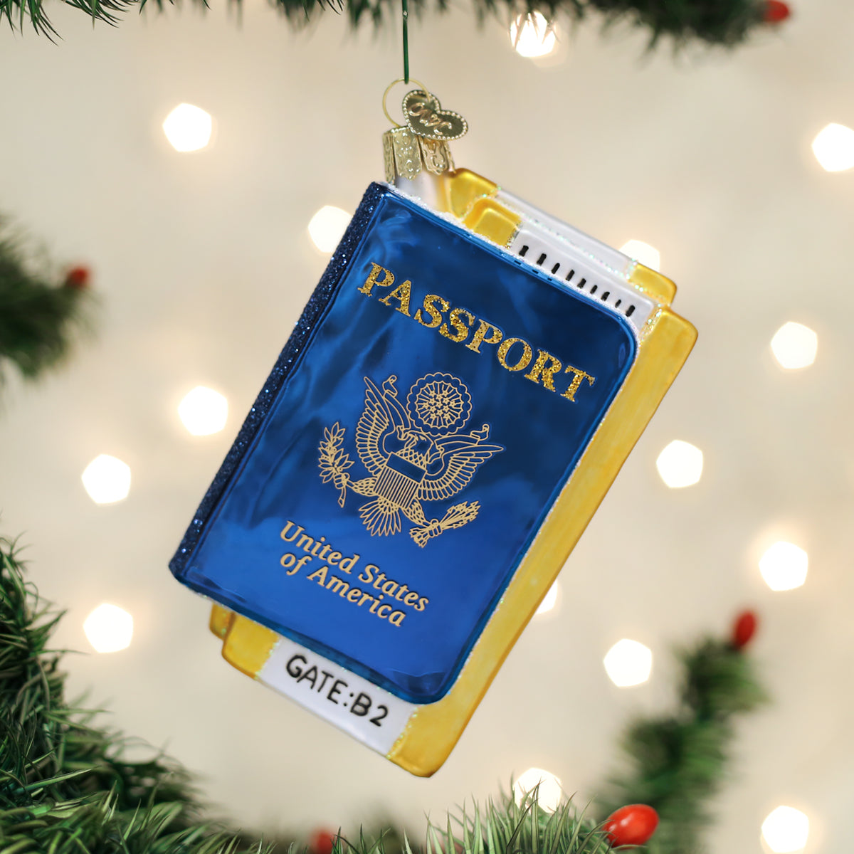 A Christmas tree ornament shaped like a blue United States passport and a yellow boarding pass labeled GATE: B2 hangs among green branches with holiday lights in the background.